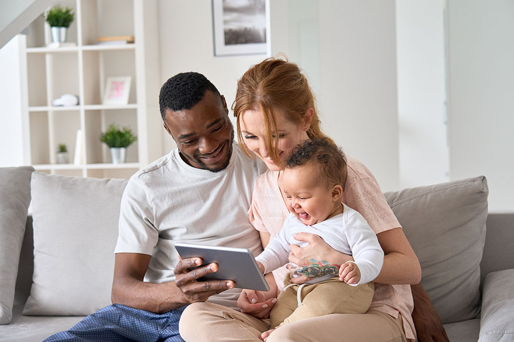 Happy young family couple holding daughter having fun with digital tablet sitting on sofa
