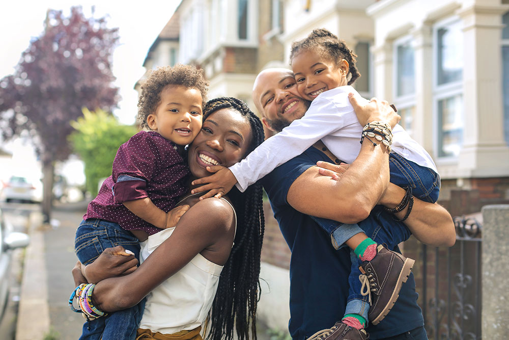 Young family holding 2 smiling children in terraced house street